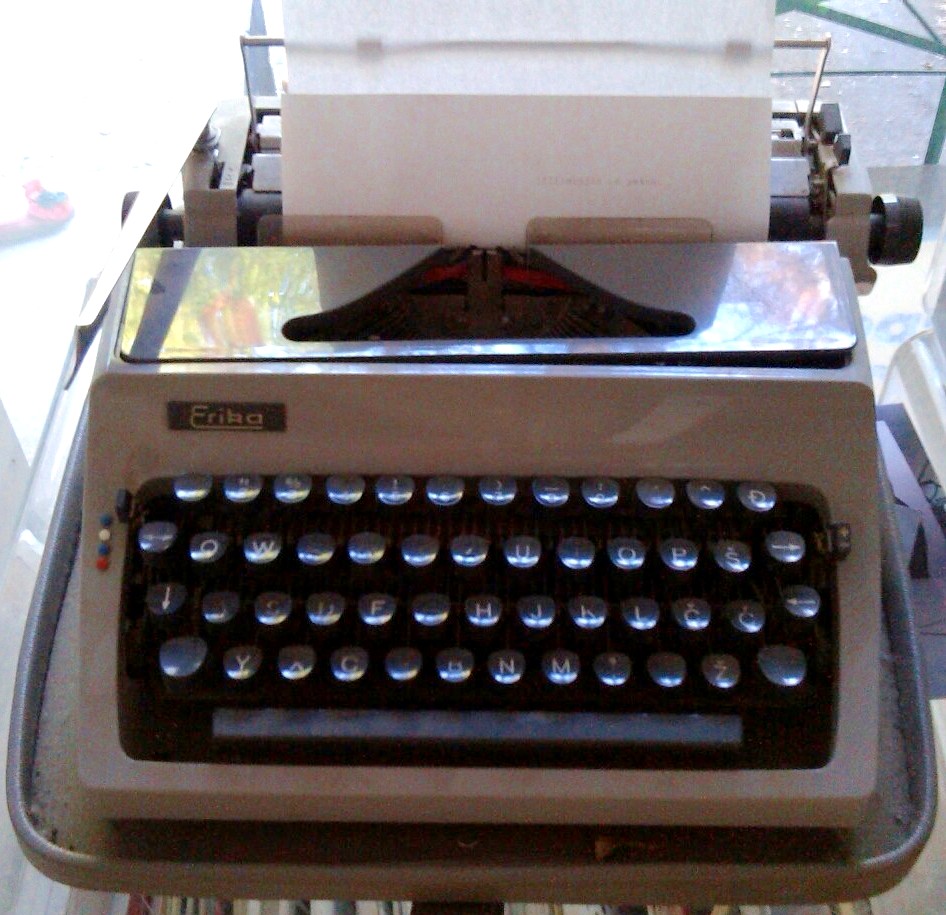 Latte-coloured typewriter with a grey keyboard and a blank piece of paper around its roller. A dark tag, front left, features the white logotype "Erika".